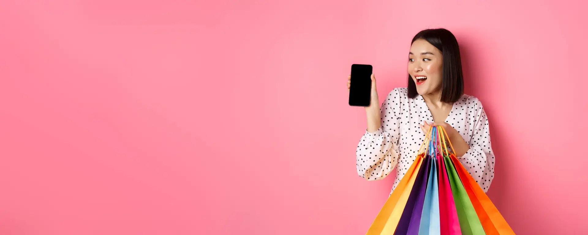 Attractive asian woman showing smartphone app and shopping bags, buying online via application, standing over pink background Autumn Delight
